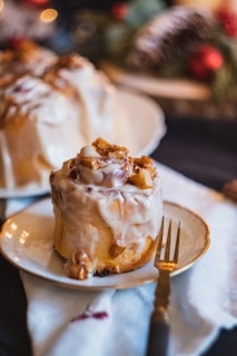 A cinnamon roll covered in creamy icing and topped with small pieces of glazed apples, placed on a white plate with a gold-rimmed fork beside it. The background features blurred decorations suggestive of a festive setting.