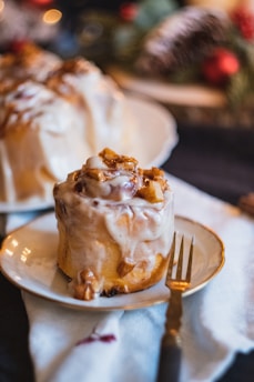 A cinnamon roll covered in creamy icing and topped with small pieces of glazed apples, placed on a white plate with a gold-rimmed fork beside it. The background features blurred decorations suggestive of a festive setting.