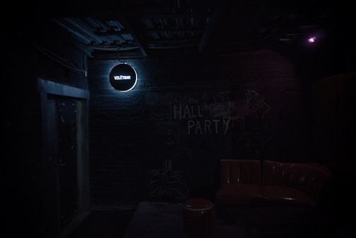 A dimly lit, industrial-style room with a dark brick wall displaying the text 'HALL PARTY' in chalk. A small, round sign with 'VZLETBAR' glows in a soft white light above a doorway. In the foreground, there is a brown leather sofa and a small round table.