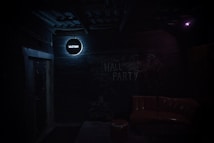 A dimly lit, industrial-style room with a dark brick wall displaying the text 'HALL PARTY' in chalk. A small, round sign with 'VZLETBAR' glows in a soft white light above a doorway. In the foreground, there is a brown leather sofa and a small round table.