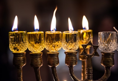 Six lit candles are placed in an ornate candelabrum made of metal with intricate designs. The candles are encased in crystal holders, and their flames cast a warm, golden light. One of the holders features a Star of David.