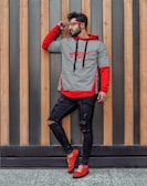 A man wearing a striped black and white shirt with a red hoodie underneath and red shoes stands against a background of vertical wooden panels. He is looking to the side and touching his sunglasses with one hand. His jeans are black and slightly torn.