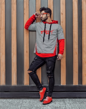 A man wearing a striped black and white shirt with a red hoodie underneath and red shoes stands against a background of vertical wooden panels. He is looking to the side and touching his sunglasses with one hand. His jeans are black and slightly torn.