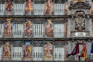 A detailed facade painted with elaborate murals depicting mythological figures and intricate decorative elements. The building features multiple windows with shutters and balconies, framed by ornate stonework. A plaque labeled 'Plaza Mayor' is visible, along with a coat of arms and flags hanging from the lower balcony.