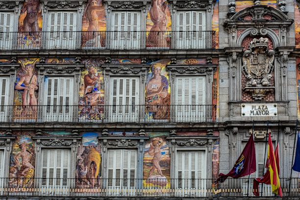 A detailed facade painted with elaborate murals depicting mythological figures and intricate decorative elements. The building features multiple windows with shutters and balconies, framed by ornate stonework. A plaque labeled 'Plaza Mayor' is visible, along with a coat of arms and flags hanging from the lower balcony.