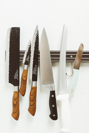 A series of kitchen knives with wooden handles are arranged on a magnetic strip against a white background. Each knife varies in size and style, and they are displayed in an orderly fashion.