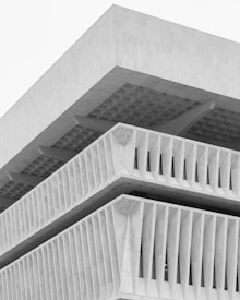 A modern architectural structure featuring geometric patterns and a focus on symmetry. The design uses sharp angles and repetitive vertical slats, creating a clean and minimalist aesthetic in a monochromatic color scheme.