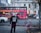 A person wearing a gas mask stands on a city street with a bold 'NEVER ENDING' sweatshirt. In the background, a vibrant pink double-decker sightseeing bus with the inscription 'CitySightseeing Berlin' is parked beside a historic building. The street is lined with bicycles attached to a rack, autumn leaves scattered on the ground, and a passing car speeds by.