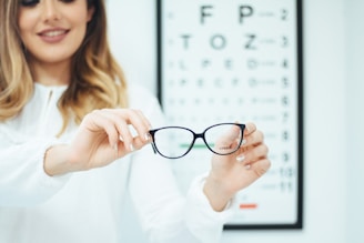 A person with long hair is holding a pair of black-rimmed glasses in both hands, slightly outstretched towards the viewer. In the background, there is an eye test chart, slightly blurred, suggesting an optometrist's setting.