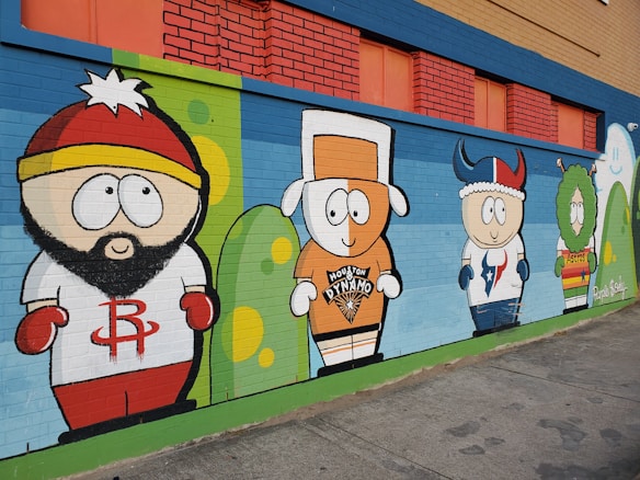 A colorful mural on a brick wall features cartoon characters wearing sports jerseys. The characters are represented in a playful, caricature style, each wearing gear from different Houston sports teams. The wall itself has a vivid background featuring blue, red, green, and yellow colors.