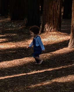 A young child wearing a long-sleeved shirt and denim overalls walks barefoot among tall trees in a forest. Sunlight filters through the trees, casting dappled shadows on the forest floor covered with dry leaves and pine needles.