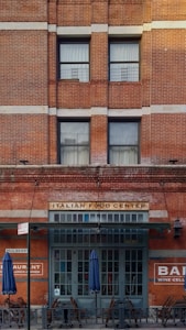 A brick building facade featuring large windows and a sign that reads 'ITALIAN FOOD CENTER' above a dark green entrance. Blue umbrellas and wooden chairs are placed outside, indicating an outdoor seating area. Additional signage on the building includes 'RESTAURANT' and 'BAR WINE CELLAR.'