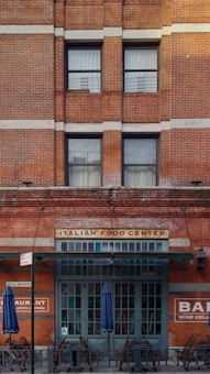 A brick building facade featuring large windows and a sign that reads 'ITALIAN FOOD CENTER' above a dark green entrance. Blue umbrellas and wooden chairs are placed outside, indicating an outdoor seating area. Additional signage on the building includes 'RESTAURANT' and 'BAR WINE CELLAR.'