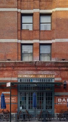 A brick building facade featuring large windows and a sign that reads 'ITALIAN FOOD CENTER' above a dark green entrance. Blue umbrellas and wooden chairs are placed outside, indicating an outdoor seating area. Additional signage on the building includes 'RESTAURANT' and 'BAR WINE CELLAR.'