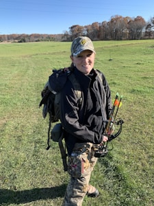 A person in outdoor attire stands in a green field under a clear blue sky. They are wearing a camouflaged cap and jacket, with a backpack and carrying a compound bow. Trees with autumn foliage are visible in the background.