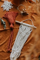 Various macramé items are arranged on a textured brown surface. One piece features intricate white knotting and fringe hanging from a wooden rod. Another is a red tassel with a thick rope design. Nearby, a delicate white lace item resembles a snowflake, and a small wreath incorporates twigs and dried flowers. A couple of leaves add a natural element to the composition.