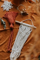 Various macramé items are arranged on a textured brown surface. One piece features intricate white knotting and fringe hanging from a wooden rod. Another is a red tassel with a thick rope design. Nearby, a delicate white lace item resembles a snowflake, and a small wreath incorporates twigs and dried flowers. A couple of leaves add a natural element to the composition.