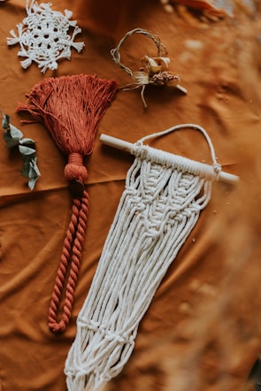Various macramé items are arranged on a textured brown surface. One piece features intricate white knotting and fringe hanging from a wooden rod. Another is a red tassel with a thick rope design. Nearby, a delicate white lace item resembles a snowflake, and a small wreath incorporates twigs and dried flowers. A couple of leaves add a natural element to the composition.