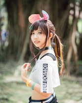 A young woman outdoors, with long hair and colorful highlights, wears a playful headband with pink and blue ears. She has a cheerful expression and is dressed in a stylish outfit with a white off-shoulder top that reads 'HAI DOMO!' in bold black letters. Wristbands in yellow and teal adorn her wrists, and she is standing in a natural setting with blurred trees in the background.