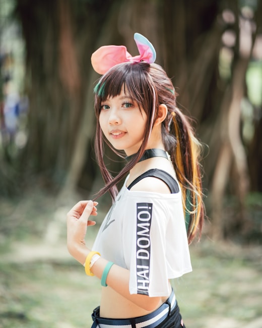 A young woman outdoors, with long hair and colorful highlights, wears a playful headband with pink and blue ears. She has a cheerful expression and is dressed in a stylish outfit with a white off-shoulder top that reads 'HAI DOMO!' in bold black letters. Wristbands in yellow and teal adorn her wrists, and she is standing in a natural setting with blurred trees in the background.