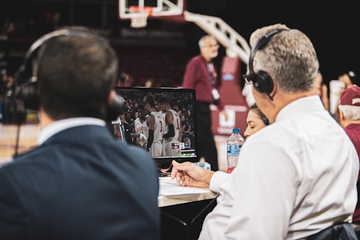 Two men wearing headsets focus on a monitor displaying a basketball team in discussion. Others can be seen around them, including a woman with a bottle of water, and an ongoing basketball game in the background.