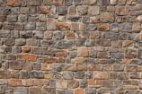 A neatly arranged stone wall with variously sized and colored rectangular stones. The stones have an uneven surface, and their colors range from shades of gray to hints of orange and brown. The mortar between the stones is visible and evenly applied.