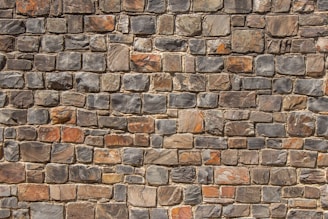 A neatly arranged stone wall with variously sized and colored rectangular stones. The stones have an uneven surface, and their colors range from shades of gray to hints of orange and brown. The mortar between the stones is visible and evenly applied.