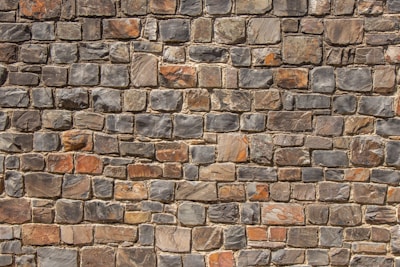 A neatly arranged stone wall with variously sized and colored rectangular stones. The stones have an uneven surface, and their colors range from shades of gray to hints of orange and brown. The mortar between the stones is visible and evenly applied.