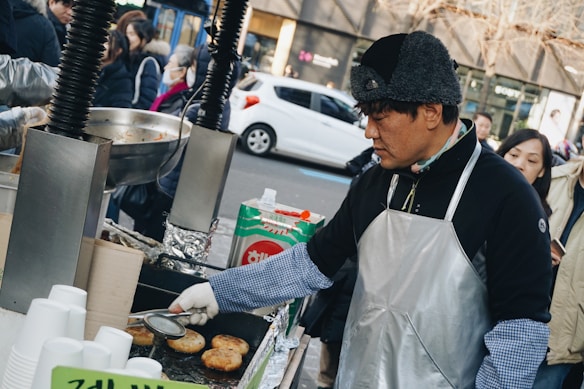 A street vendor is cooking food at an outdoor stall, wearing a silver apron and a winter hat. The vendor appears focused on using a spatula to turn the food items on a flat grill. Several people are standing nearby, some wearing winter clothing. A stack of white disposable cups is visible, and in the background, a white car is parked on the street.