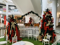 A festive indoor display featuring two decorated Christmas trees adorned with red and gold ribbons. Two reindeer figures are positioned between the trees, bordered by a white picket fence. Several white chairs encircle a round table with a coloring book on it, all situated on green artificial grass. The setting appears to be a shopping mall, with visible railings and storefronts in the background.
