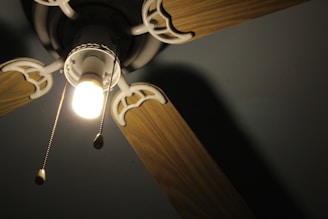 An overhead view of a ceiling fan with wooden blades and a central light fixture. The light is turned on, emitting a warm glow, and two pull chains are visible hanging from the fixture.