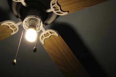 An overhead view of a ceiling fan with wooden blades and a central light fixture. The light is turned on, emitting a warm glow, and two pull chains are visible hanging from the fixture.