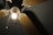 An overhead view of a ceiling fan with wooden blades and a central light fixture. The light is turned on, emitting a warm glow, and two pull chains are visible hanging from the fixture.