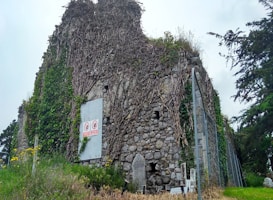 An old stone building overgrown with vines and ivy, featuring weathered walls and a metal sign with prohibition symbols. The scene includes a few gravestones in front and trees in the background, creating a setting that feels both natural and historical.