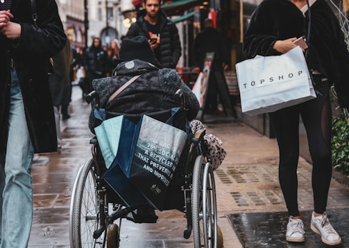 A busy street scene with a person in a wheelchair carrying several shopping bags. Other pedestrians walk alongside, some carrying more bags. The ground is wet, suggesting recent rain, and the area is lined with shops.