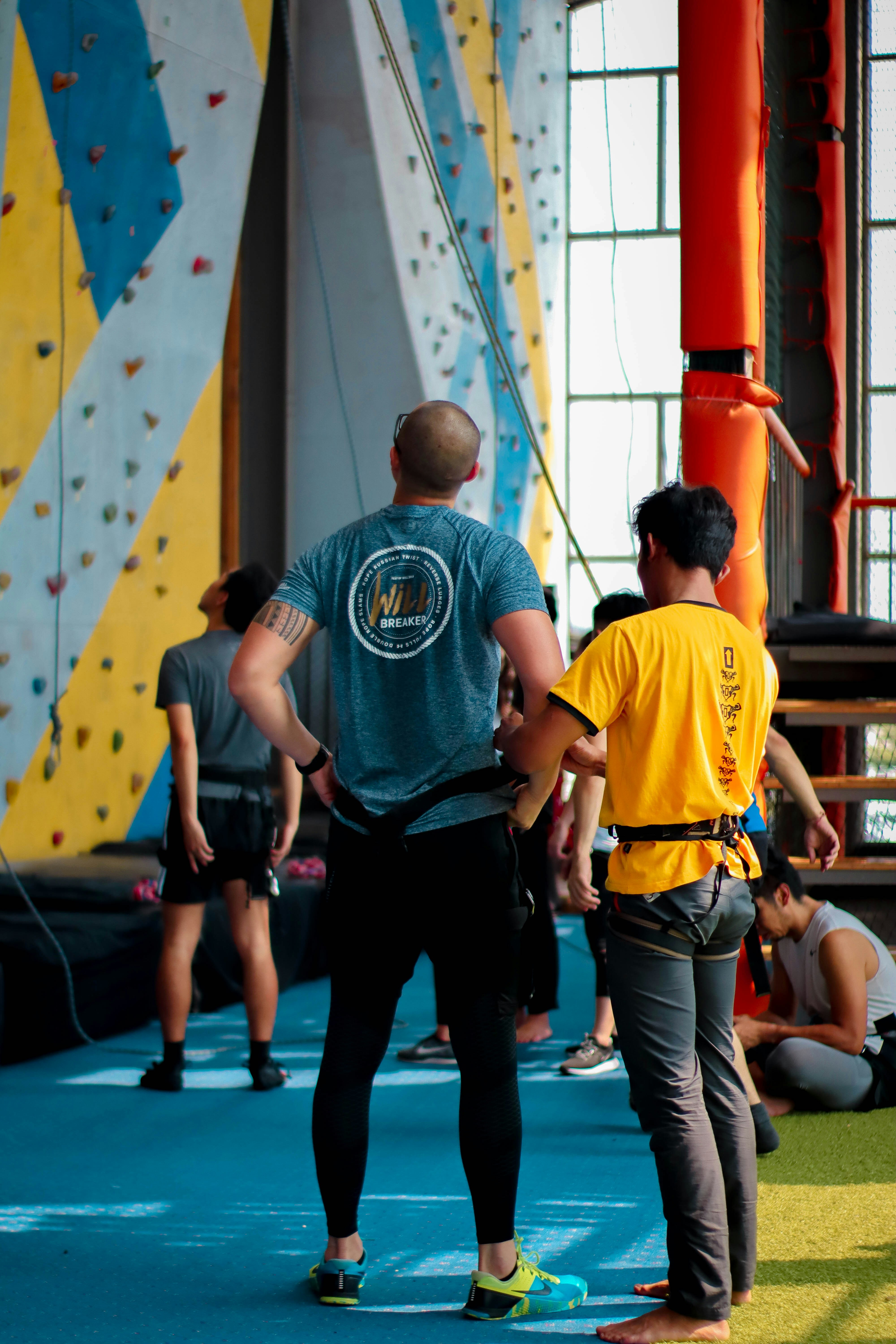 Several people are standing in an indoor rock climbing gym. Two individuals in the foreground are wearing climbing gear and are looking towards the climbing wall. The walls are covered with multicolored climbing holds. There is an orange structural pole, and a blue cushioned floor covering the area.