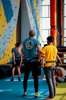 Several people are standing in an indoor rock climbing gym. Two individuals in the foreground are wearing climbing gear and are looking towards the climbing wall. The walls are covered with multicolored climbing holds. There is an orange structural pole, and a blue cushioned floor covering the area.