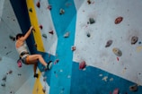 A person is climbing an indoor rock wall with various colored holds. The wall features a dynamic design with blue, yellow, and white sections. The climber is wearing a tank top, shorts, climbing shoes, and has a chalk bag attached to their waist.