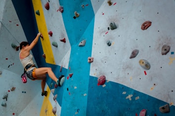 A person is climbing an indoor rock wall with various colored holds. The wall features a dynamic design with blue, yellow, and white sections. The climber is wearing a tank top, shorts, climbing shoes, and has a chalk bag attached to their waist.