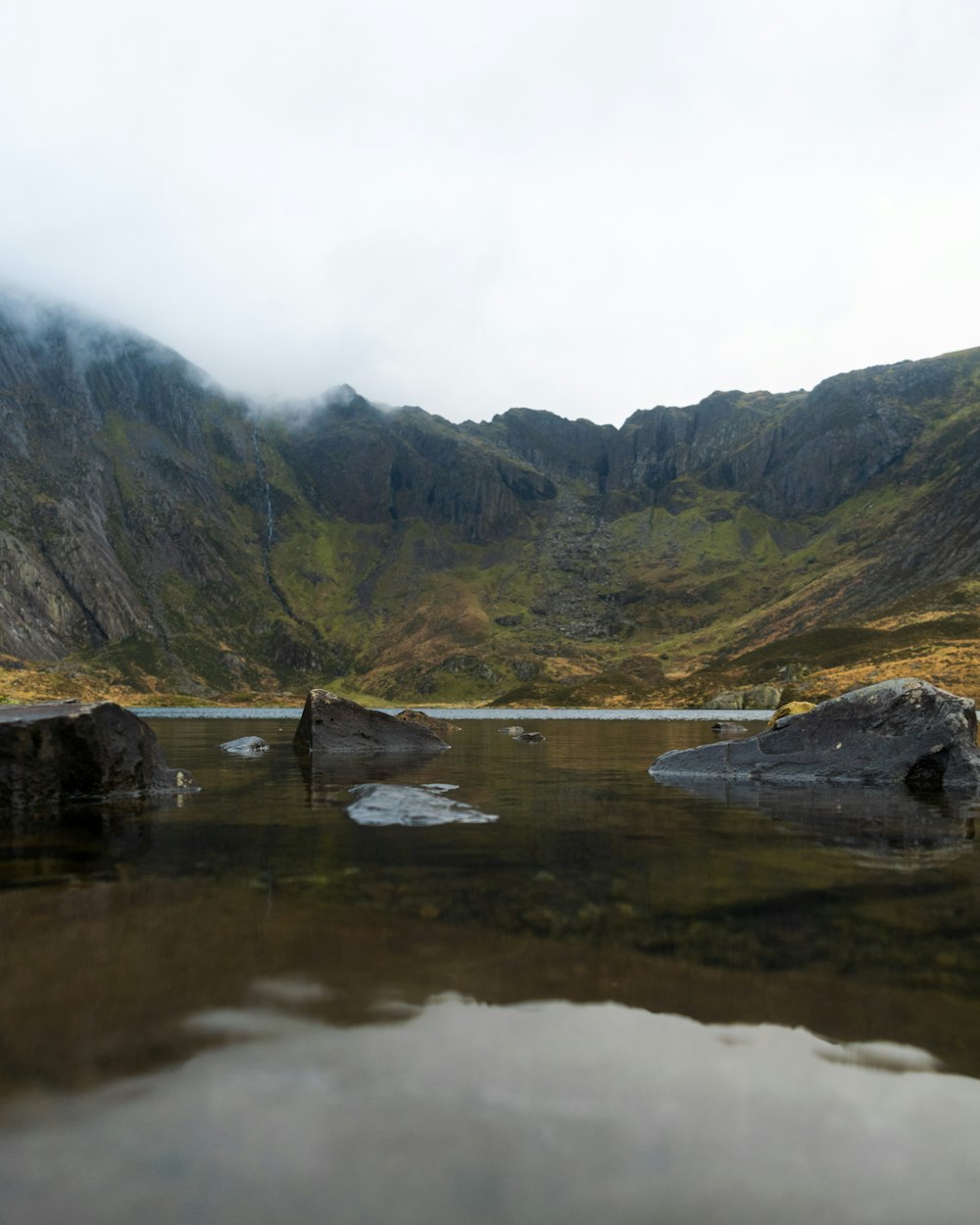 gray rock formations on body of water viewing green field and mountain under white sky