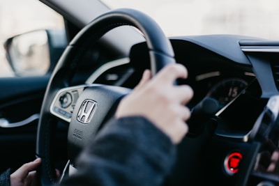 A person is gripping the steering wheel of a Honda vehicle. The focus is on the steering wheel, with the Honda logo prominently displayed. The car's interior features modern design elements, including a digital dashboard and control panel.