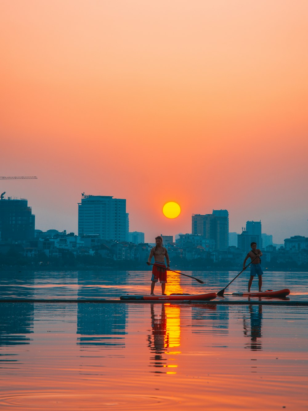 two topless men standing on paddle board on body of water viewing city with high-rise buildings under orange sky