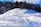 A group of people are trekking through a snowy landscape, casting long shadows on the white surface. The snow-covered hills in the foreground lead up to a forest of evergreen trees under a bright blue sky. The sunlight enhances the textures of the snow and adds depth to the shadows.
