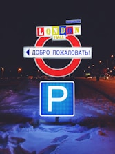 A parking sign is illuminated at night, surrounded by snow. Above it, a sign reads 'LONDON MALL' in bright, colorful letters, and a Russian sign welcomes visitors.