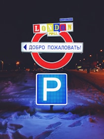 A parking sign is illuminated at night, surrounded by snow. Above it, a sign reads 'LONDON MALL' in bright, colorful letters, and a Russian sign welcomes visitors.