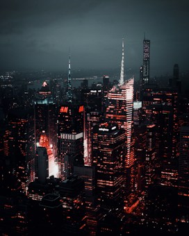 A nighttime cityscape showcasing towering skyscrapers illuminated with vibrant red and white lights against a dark, cloudy sky. The buildings are densely packed and the ambient glow highlights the architectural features and logos, creating a striking contrast with the surrounding darkness.