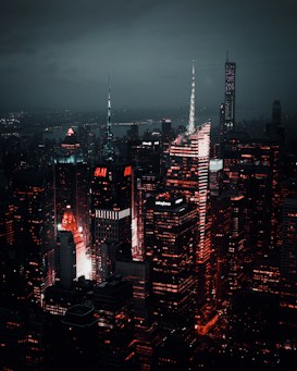 A nighttime cityscape showcasing towering skyscrapers illuminated with vibrant red and white lights against a dark, cloudy sky. The buildings are densely packed and the ambient glow highlights the architectural features and logos, creating a striking contrast with the surrounding darkness.