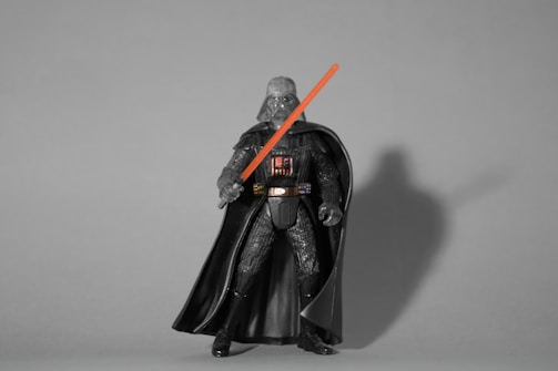 A monochrome action figure dressed in a detailed, dark outfit with a long flowing cape holds a glowing red light sword. The figure stands against a plain gray background with a distinct shadow cast to one side.
