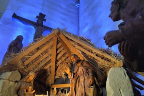 A nativity scene featuring statues of Mary, Joseph, and baby Jesus inside a thatched roof stable. In the background, there is a crucifix with a figure of Jesus on the cross, illuminated by blue lighting. The scene includes hay and wooden beams, enhancing the rustic and sacred atmosphere of the setting.