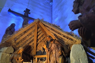 A nativity scene featuring statues of Mary, Joseph, and baby Jesus inside a thatched roof stable. In the background, there is a crucifix with a figure of Jesus on the cross, illuminated by blue lighting. The scene includes hay and wooden beams, enhancing the rustic and sacred atmosphere of the setting.